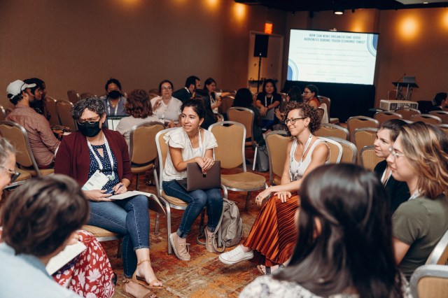 A group of about 10 conference attendees gather in a circle to discuss a prompt question, displayed on the screen in the background: How can news organizations engage audiences during tough economic times?
