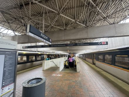 The Atlanta Airport MARTA Station stop platform is nearly empty. There is an overhead sign indicating directions to the airport and other destinations. An escalator in the center leads down to the train tracks, where a few people are boarding stationary trains on either side. The ceiling features a lattice of metal beams.