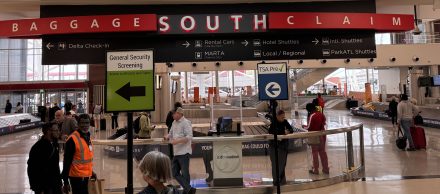 Wide-angle view of a busy airport baggage claim area with various signs, including "Baggage South Claim," "General Security Screening," and "TSA Pre-check." People are walking or waiting, with baggage carousels, rental car counters, and hotel shuttle services in the background.