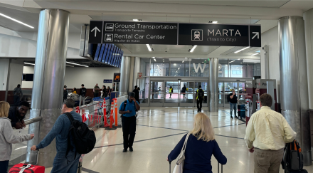 Travelers walk through an airport corridor, with baggage claim to the left, under a sign directing them to ground transportation, rental car center, and MARTA (train to city). The sign includes Spanish translations, and the environment appears busy with passengers and security personnel.